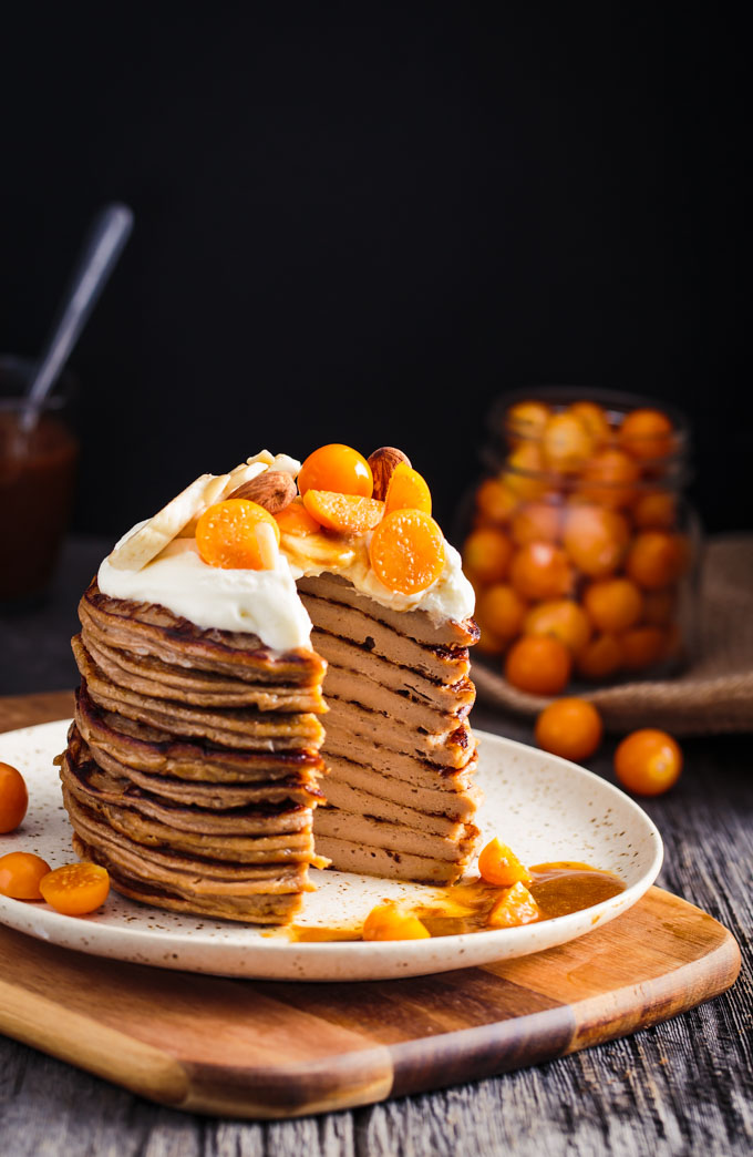 A stack of pancakes with whipped cream and fruits on top. A triangle slice is cut out.